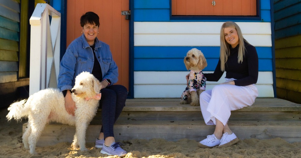 two women sitting outside a beach hut with their dogs