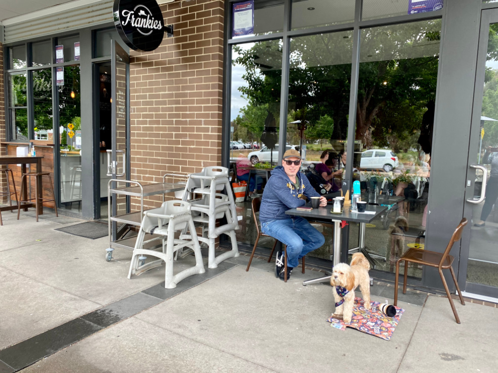 man sitting at a table with coffee and cavoodle sitting underneath