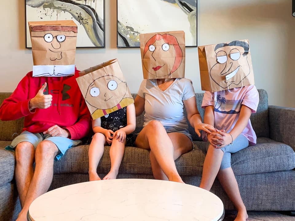 family of four sitting on sofa wearing paper bags decorated with funny faces over their heads