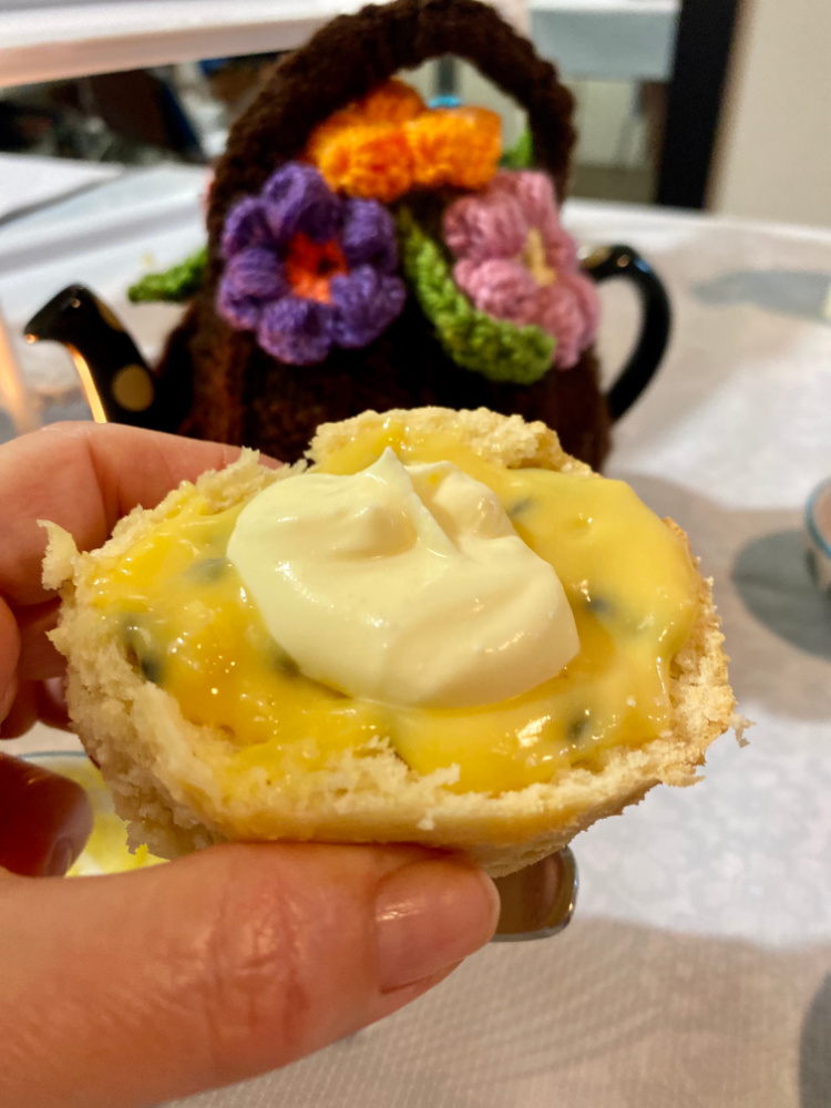 hand holding scone with passionfruit curd and cream and teapot with tea cosy in background