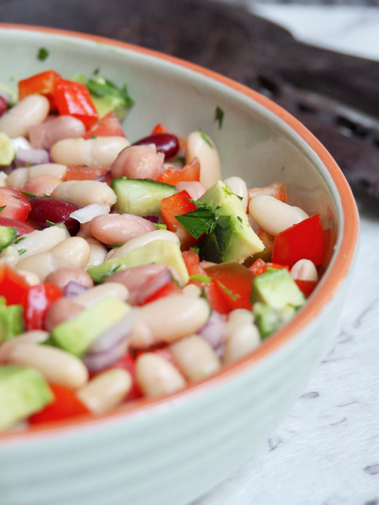 light green dish full of mixed bean salad with chopped tomatoes avocado cucumber red onion and mixed beans