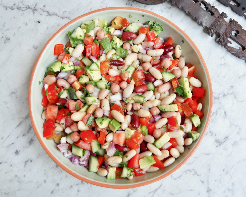 light green dish full of mixed bean salad with chopped tomatoes avocado cucumber red onion and mixed beans