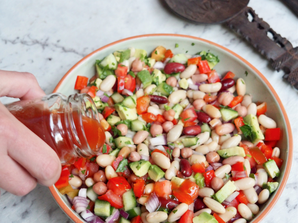 hand holding jar pouring dressing over mixed bean salad with chopped tomatoes avocado cucumber red onion and mixed beans