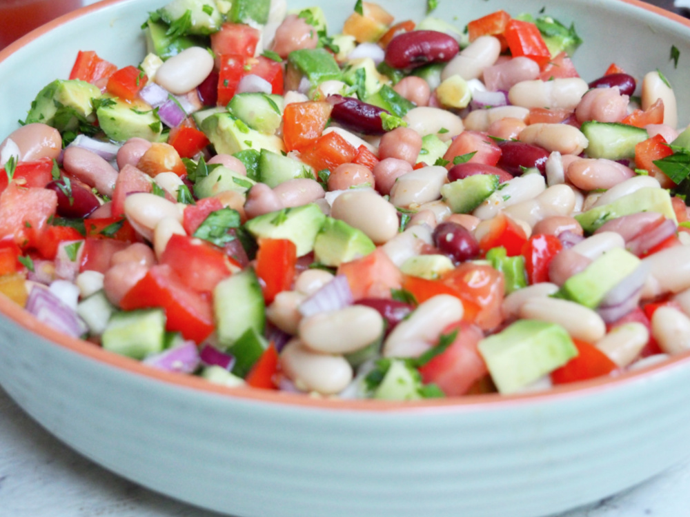 light green dish full of mixed bean salad with chopped tomatoes avocado cucumber red onion and mixed beans