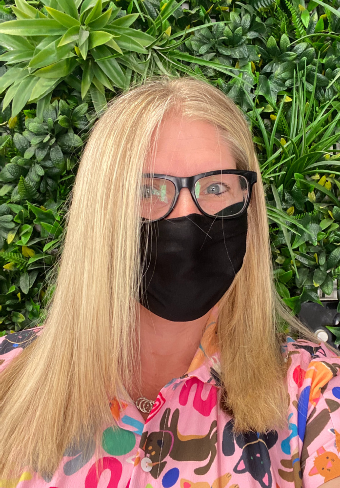woman wearing black glasses with black face mask and freshly cut long blonde hair standing in front of a plant wall