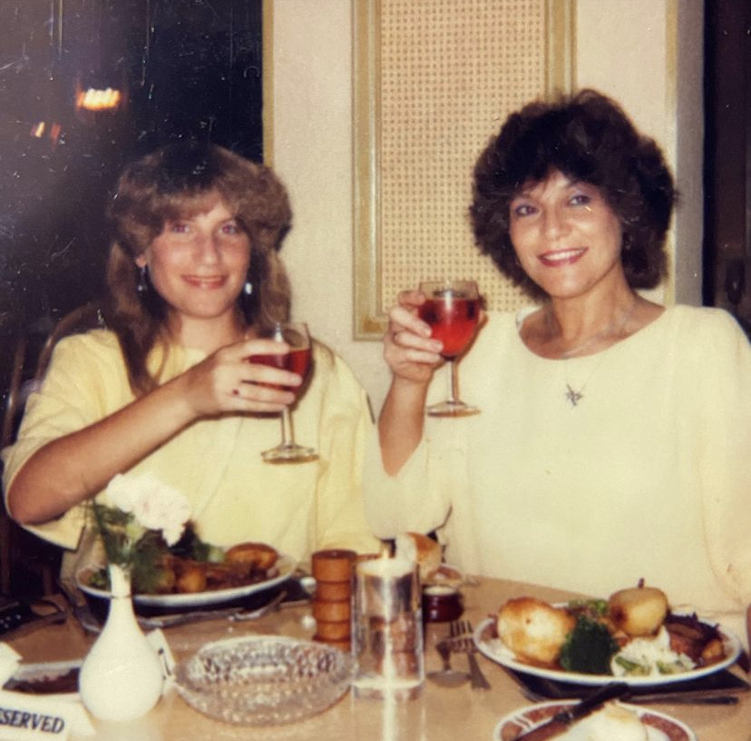 girl with permed hair and lady with brown wavy hair both wearing yellow dresses and toasting a glass of red wine at a restaurant table