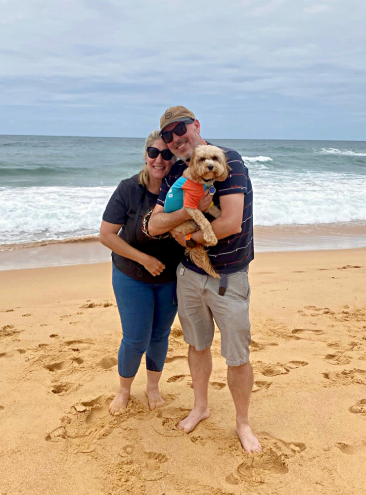 a man and a woman standing close together on a beach on a cloudy day with the ocean in the background. The man is holding a cavoodle wearing a lifesaver rashie.