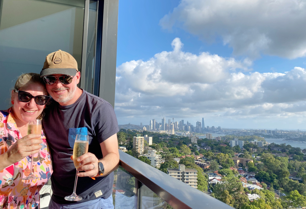 a man and woman holding flutes of champagne on balcony with view of sydney in background