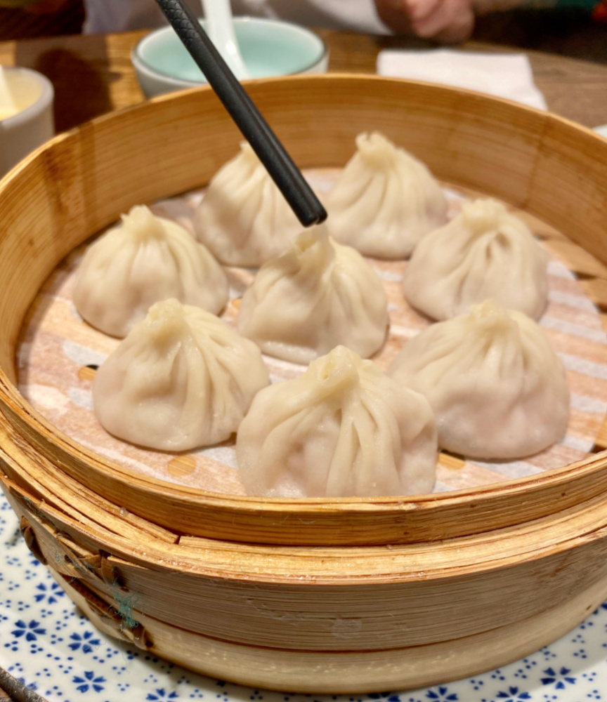 a bamboo steamer full of xiao long bao with a pair of chopsticks above touching one of the dumplings
