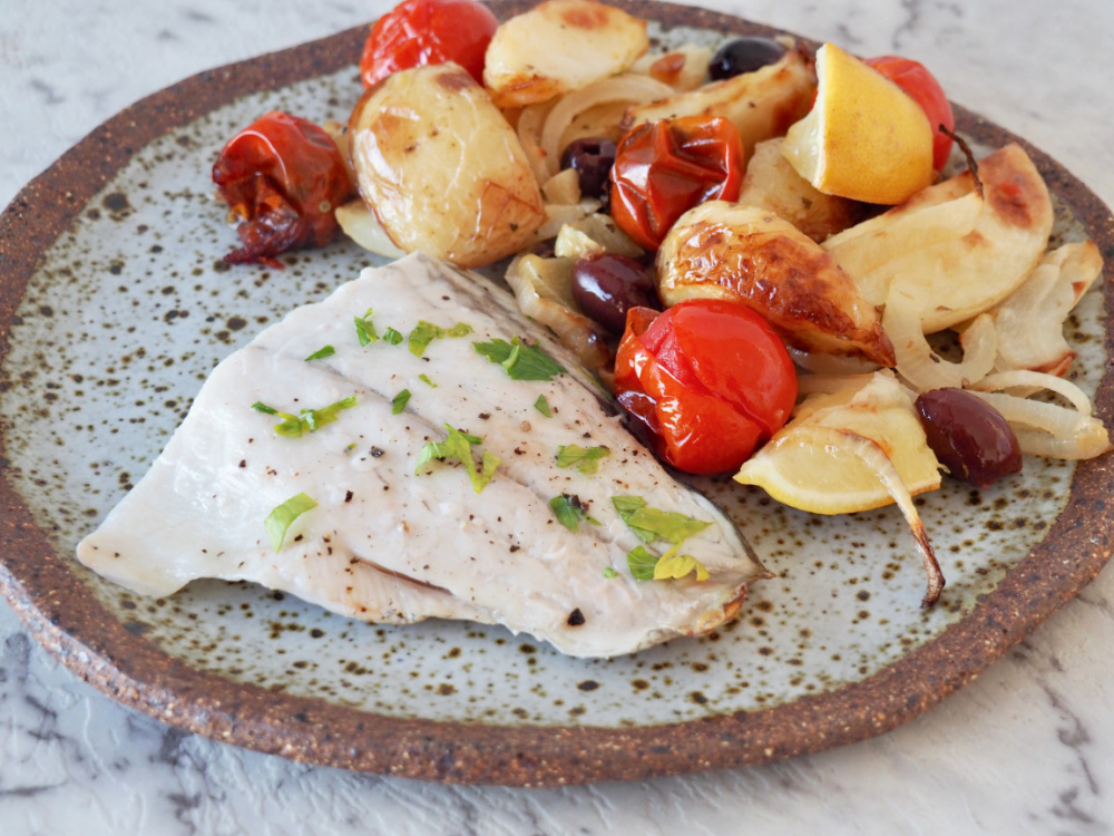 white fish garnished with parsley on stone plate with roasted potatoes, tomatoes and onions