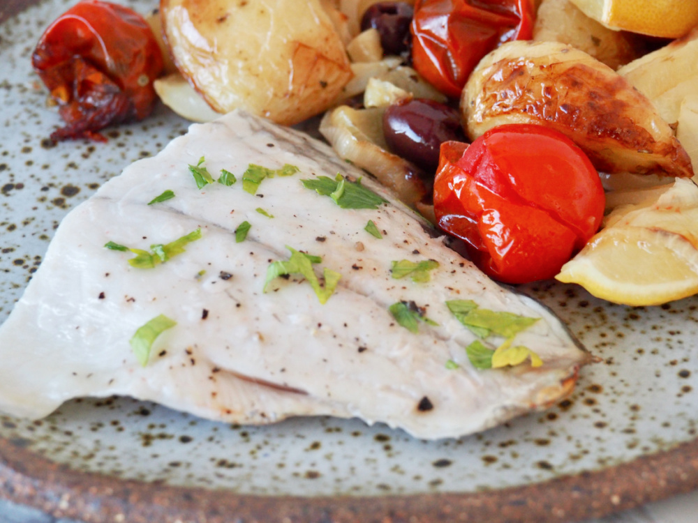 white fish garnished with parsley on stone plate with roasted potatoes, tomatoes and onions