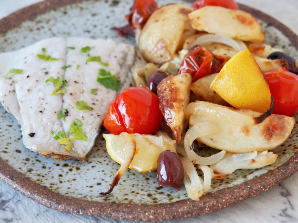 white fish garnished with parsley on stone plate with roasted potatoes, tomatoes and onions