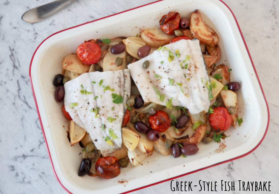 two fish fillets on a bed of roasted sliced onion, tomatoes, olives, capers and potato wedges in a small white enamel baking tin