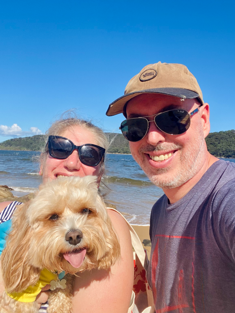 selfie of a man and a woman, the woman is holding a cavoodle who is looking at the camera. They are on a beach and the ocean is in the background