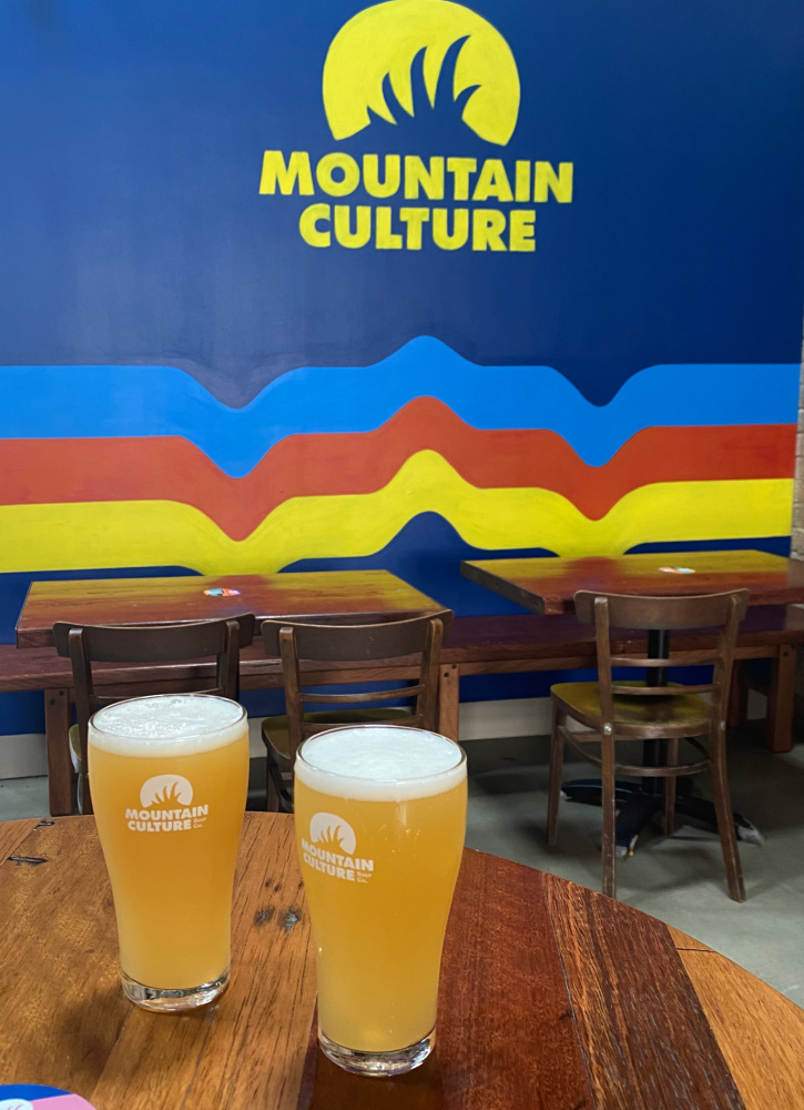 2 pints of mountain culture beer in front of a blue wall with the words Mountain Culture in block yellow capitals