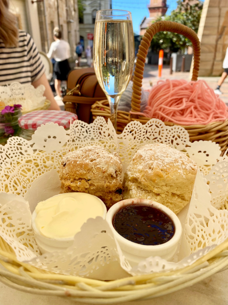 2 scones a pot of cream and a pot of jam on plate with paper doillie and glass of champagne in the background