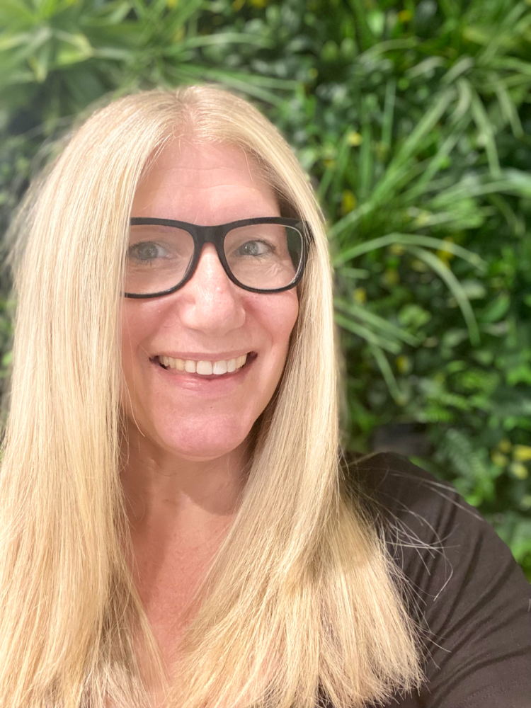 woman with long blonde hair and glasses smiling in front of a plant wall