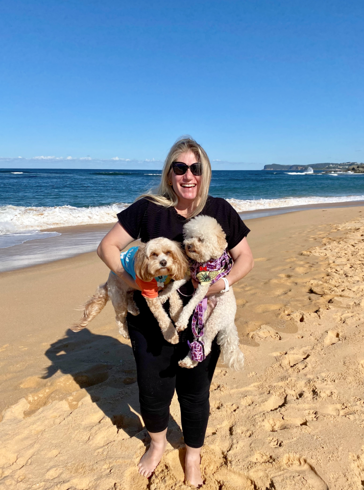 woman wearing black top and black leggings is standing on a beach with the ocean behind her and is holding a small fluffy dog in each arm