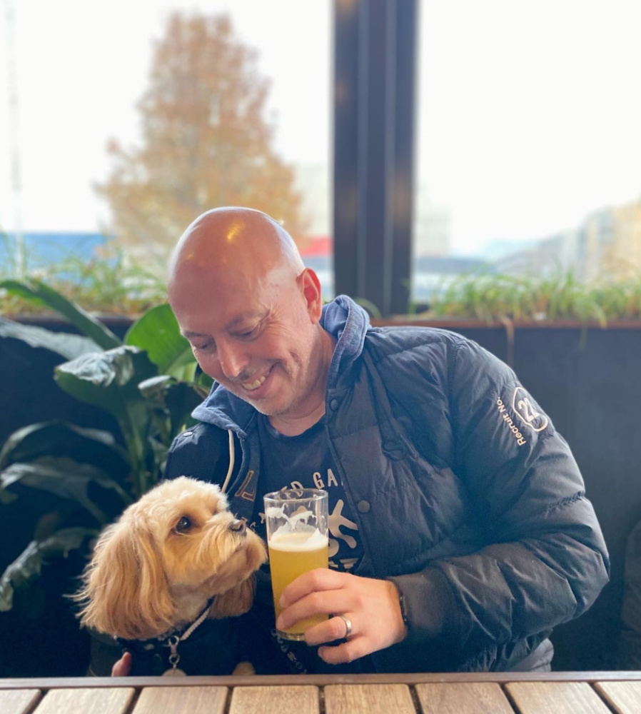 a man is sitting next to a cavoodle who is looking lovingly at the glass of beer he is holding