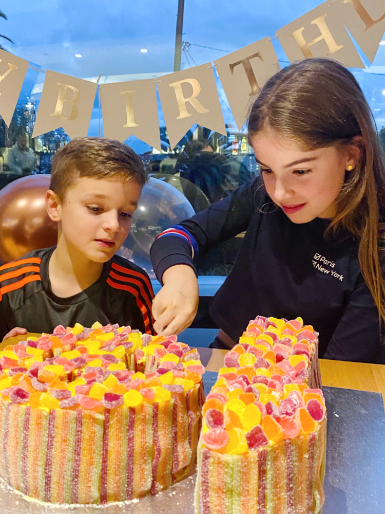 a boy and a girl are looking excitedly at a number 10 cake covered in coloured sweets. The girl is cutting into the cake and smiling.