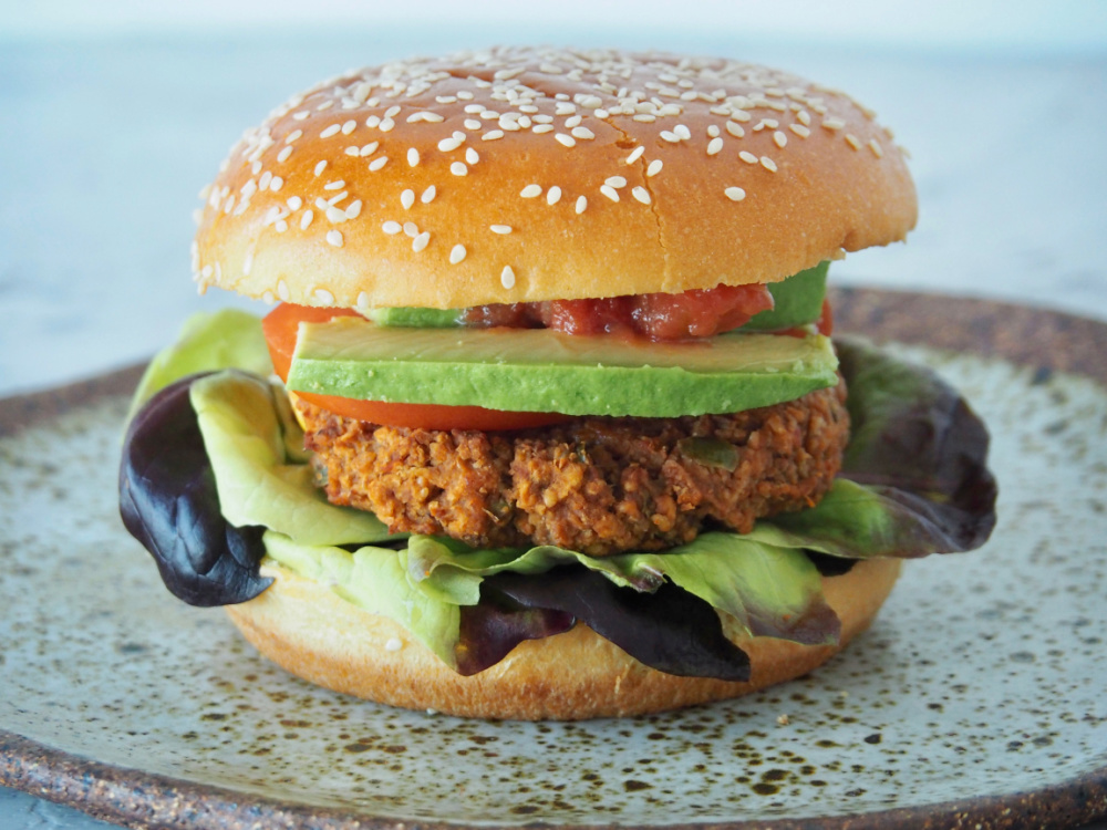 refried bean burger stacked in a sesame seed bun with red oak lettuce, sliced avocado and salsa