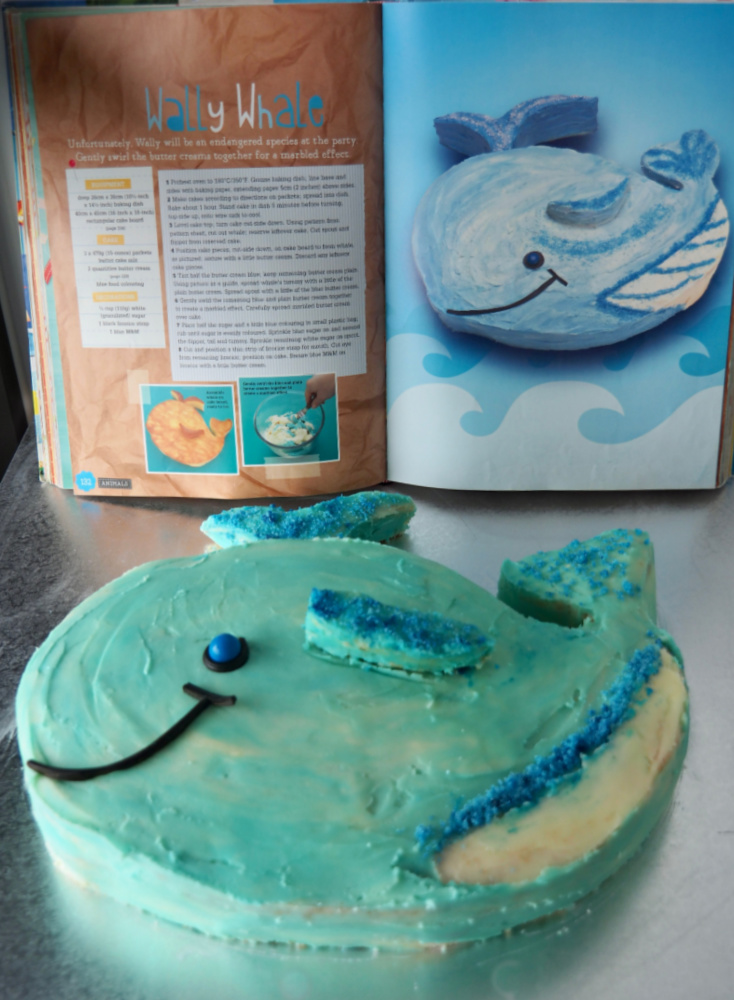 whale cake on a silver board with recipe book showing how to make in background