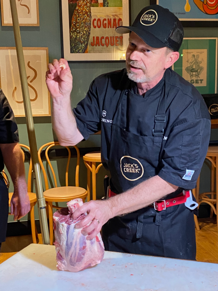 a butcher in a black apron and black cap holds a hammer steak in one hand and a small piece of marrow from the bone in the other