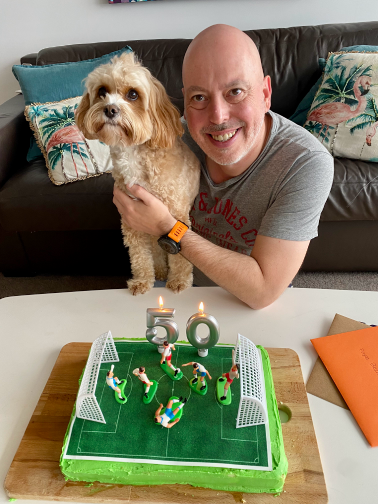 a smiling man is holding a cavoodle and sitting behind a low coffee table with a soccer pitch birthday cake on it