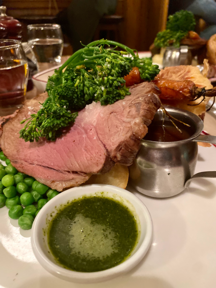 big slices of roast lamb, broccolini and roast potatoes on a plate with a pot of mint sauce at the front