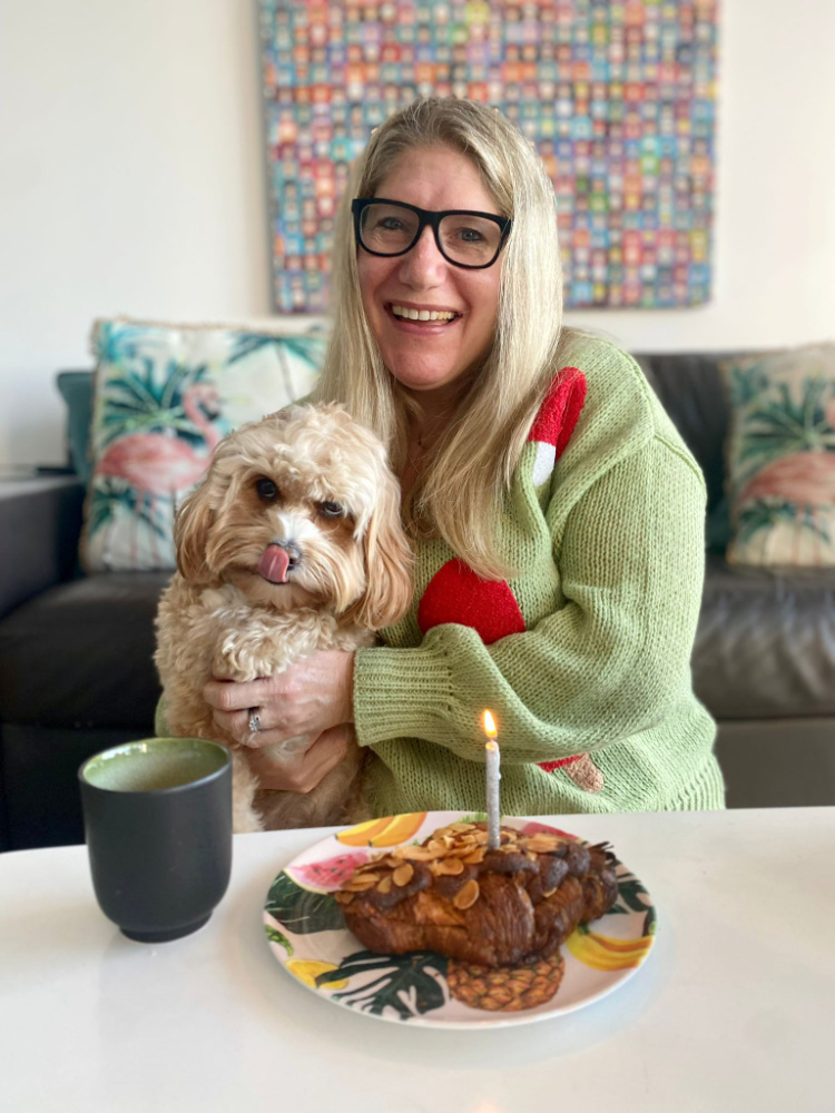 a woman with long blonde hair holding an apricot coloured cavapoo looking at a croissant with a candle placed on a low coffee table