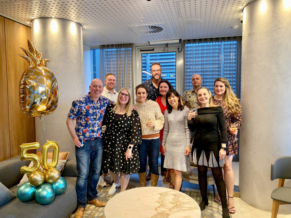 a group of 10 people smiling and standing next to a large pineapple balloon and balloons saying the number 50