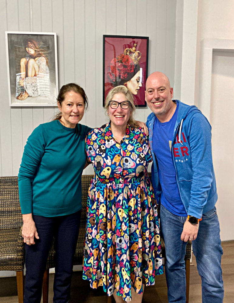 Thee people standing smiling at the camera in front of a wood panelled wall with two portraits