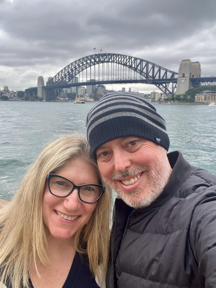 a selfie of woman with blonde hair and glasses and a man with a beard and a beanie in front of the Sydney Harbour Bridge