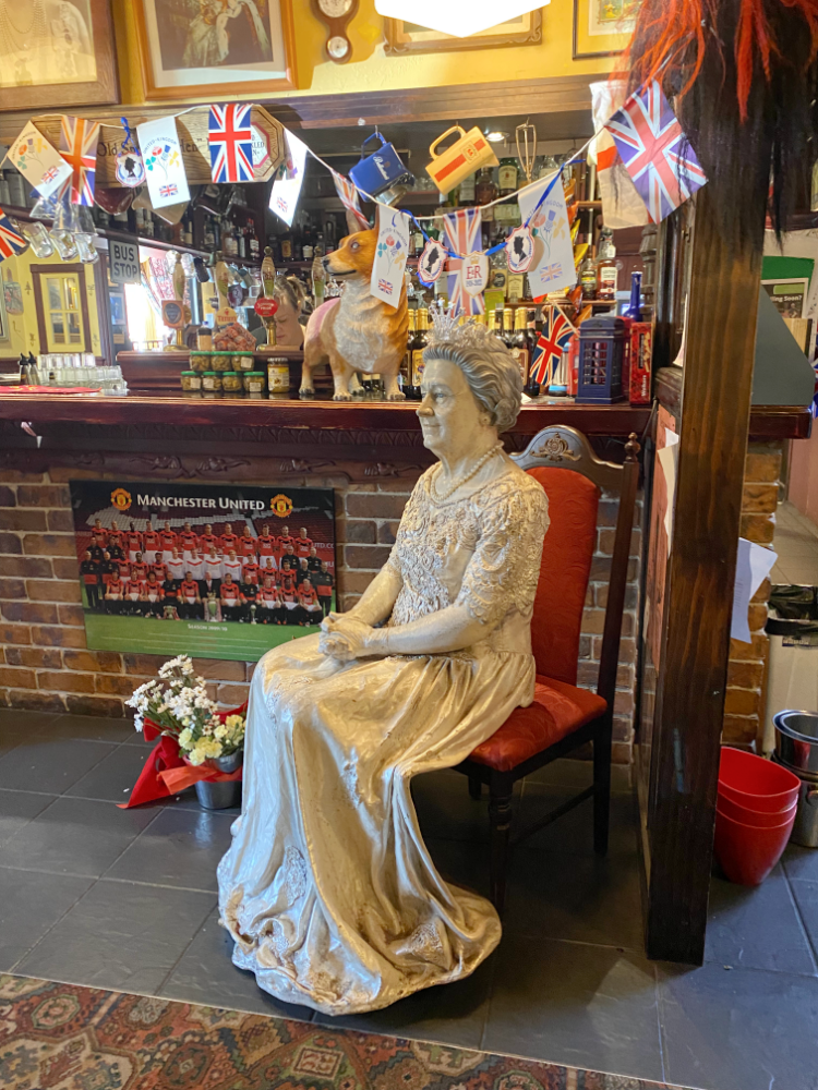 a statue of the Queen sitting down in front of a  bar decorated with all kinds of memorabilia