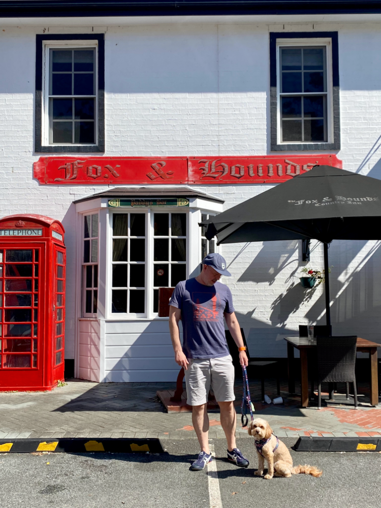 a man looking down at a cavapoo outside a pub with white walls next to a red phone box