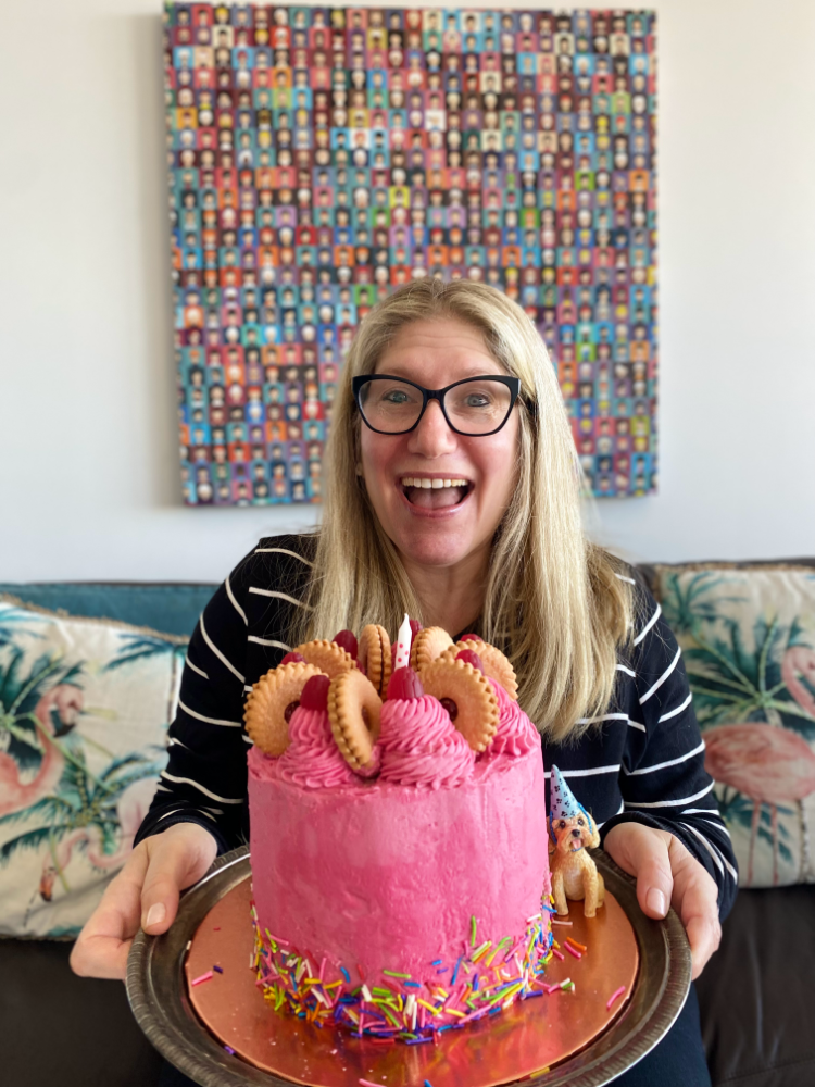 woman wearing black glasses and smiling widely holding a bright pink cake topped with pink swirls and jammy dodger biscuits