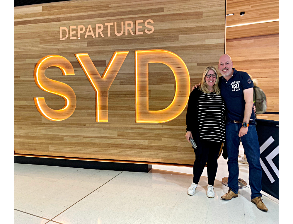 a man and woman wearing black tops and leggings and jeans standing by the SYD sign at Sydney airport