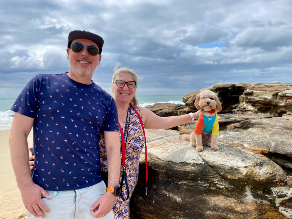 A man and woman standing next to a rock. A little cavoodle is sitting on the rock and the woman is stretching over and patting him on the back.