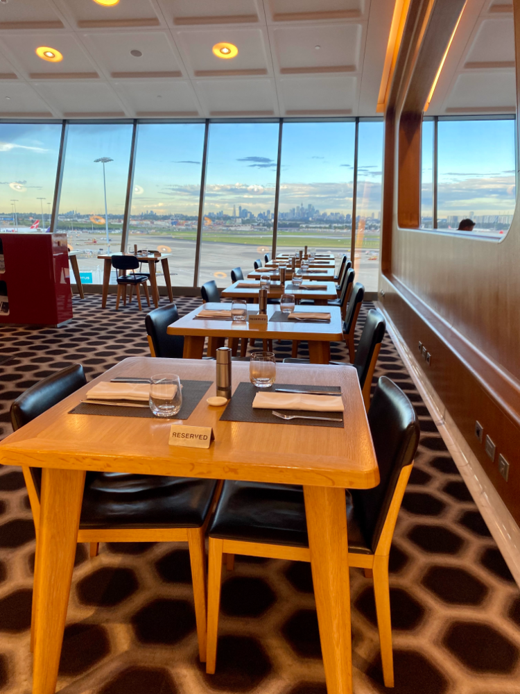 looking at the dining section of the first class lounge with a row of laid wooden tables looking out on to the airport runway