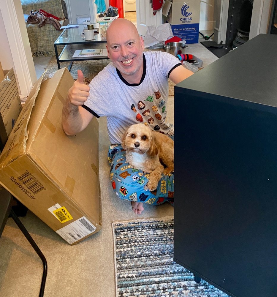 A man wearing star wars pyjamas sitting with a dog on his lap assembling flat pack furniture