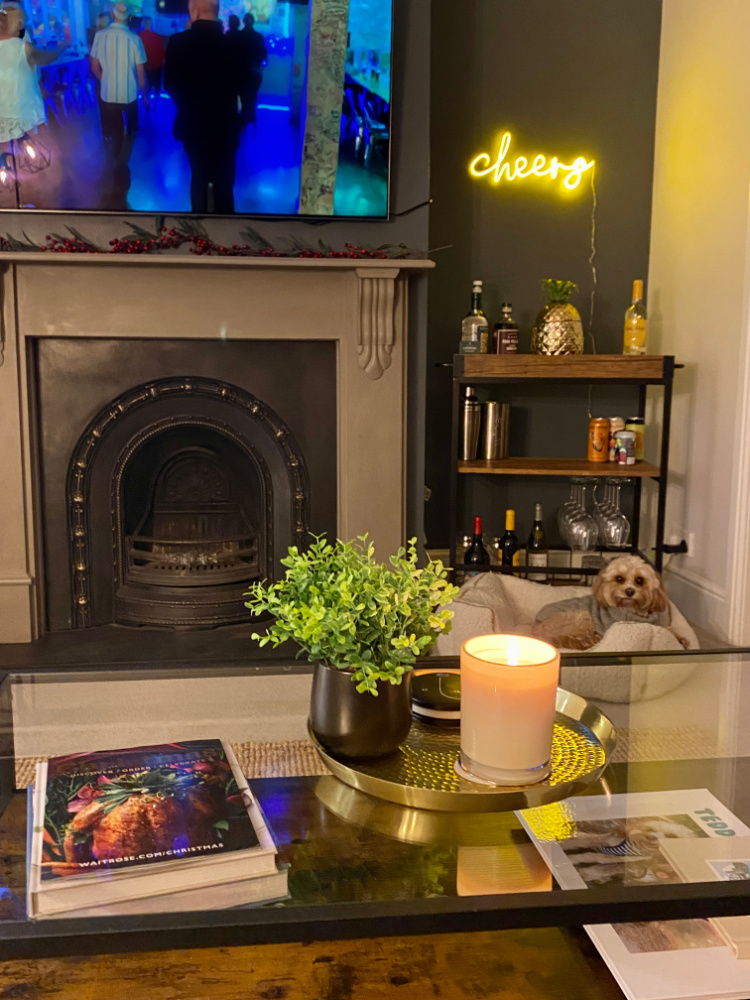 living room scene with a dog lying in a bed in front of a fireplace. There is a bar cart next to the fireplace and above it is a neon yellow sign saying Cheers