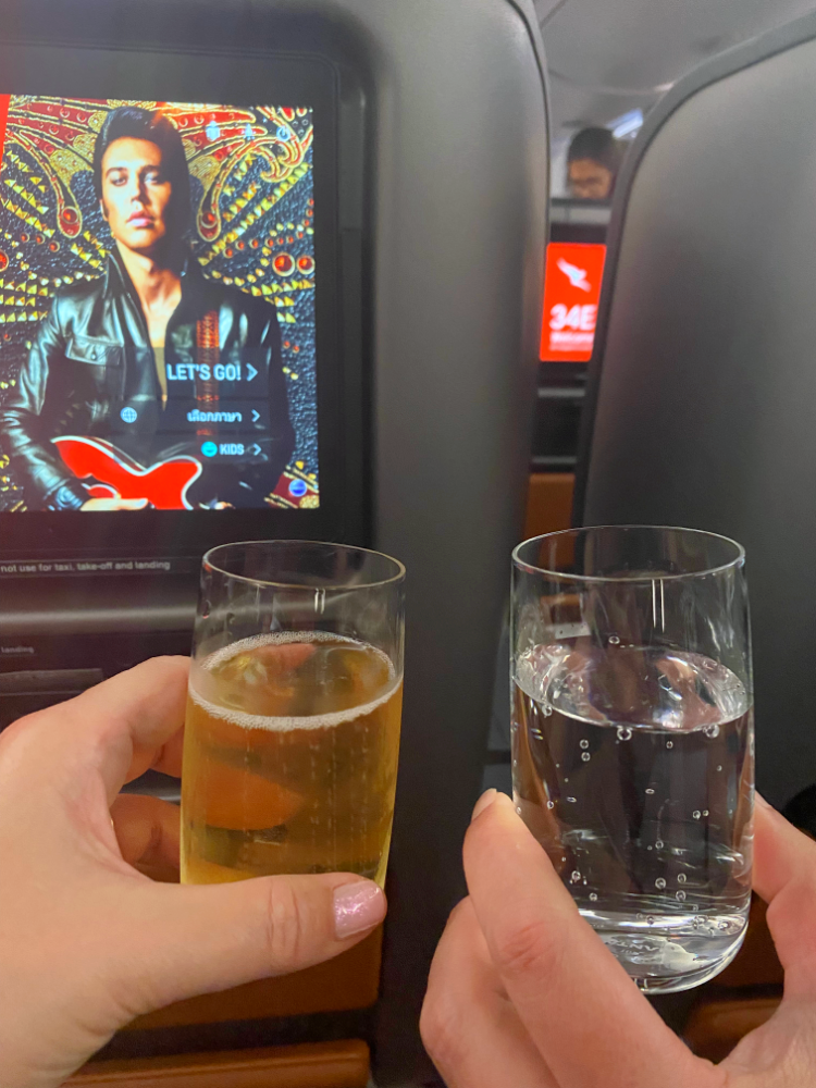 a glass of bubbles and a glass of sparkling water being raised in front of a seat screen on a plane