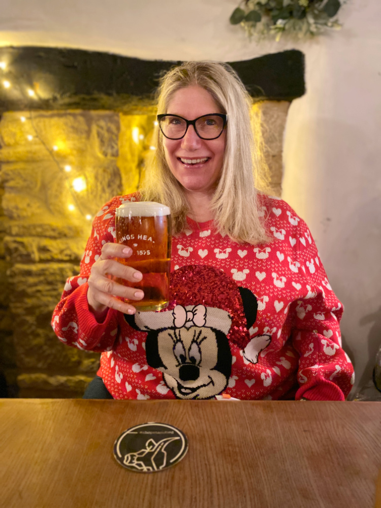 woman with blonde hair and glasses wearing a red minnie mouse christmas jumper and raising a pint of beer