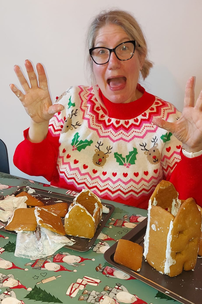 woman wearing christmas jumper with hands in air looking shocked with a destroyed gingerbread house in front of her