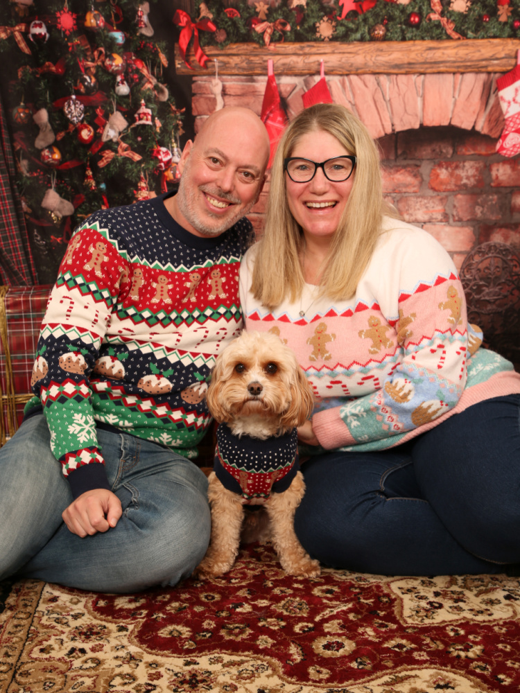 a man, woman and a small cavapoo sitting on the floor in front of a christmas backdrop wearing matching christmas jumpers