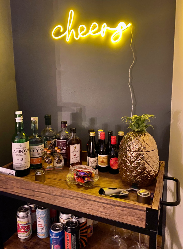 A bar trolley stacked full of bottles and a big gold pineapple ice bucket. Above the bar trolley is a yellow neon sign that says Cheers.