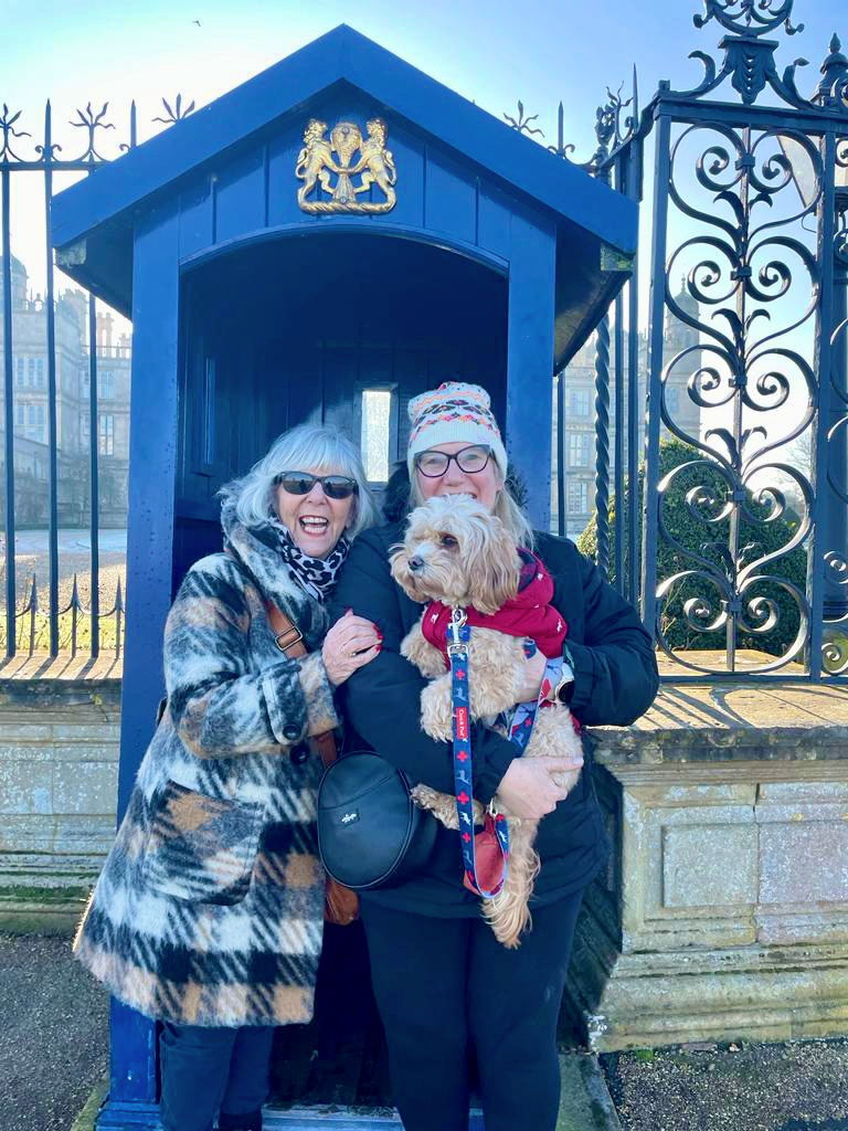 a woman with a checked coat and grey hair cuddling up to a woman with a bobble hat holding a small cavapoo wearing a red coat