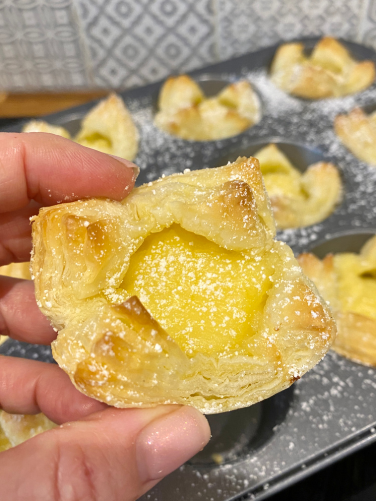 a hand holding a small puff pastry puff filled with lime curd. In the background there is a tray of lime puffs.