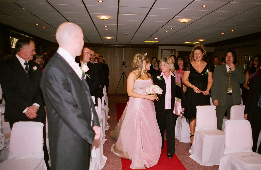 woman in suit walking next to woman wearing a pink wedding dress down the aisle
