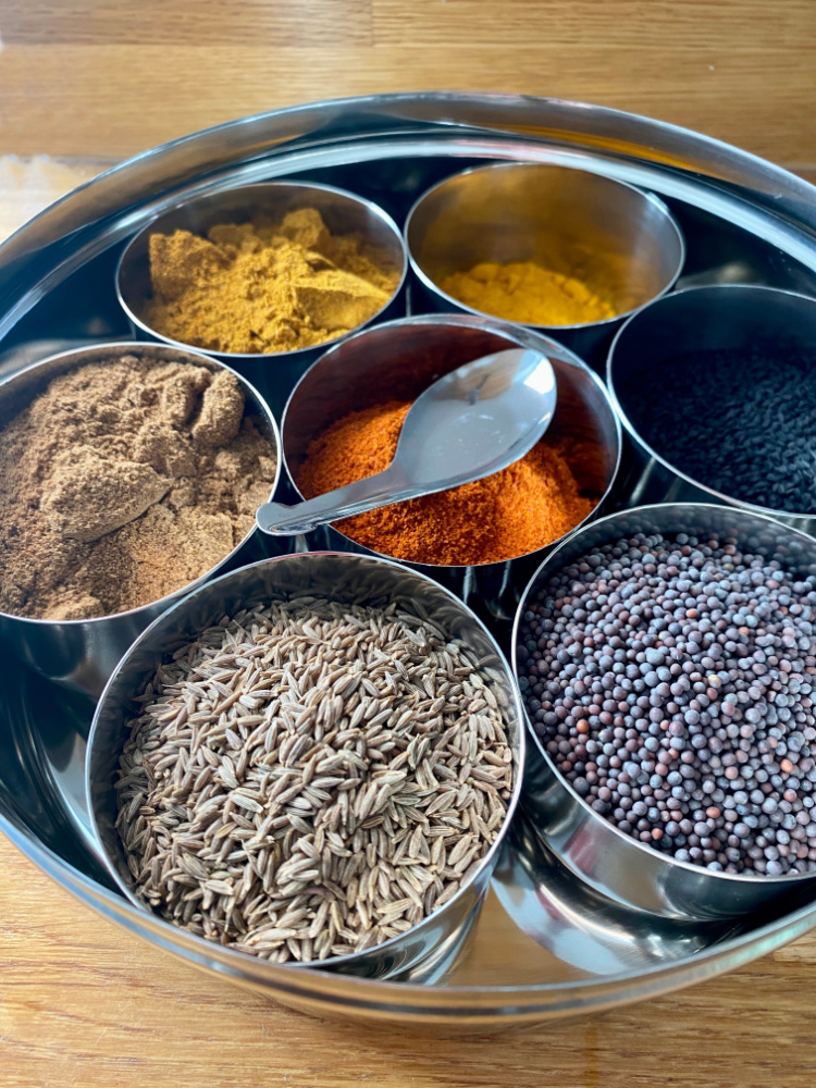 a stainless steel spice tin with 7 small bowls filled with spices. There is a small teaspoon resting on the top